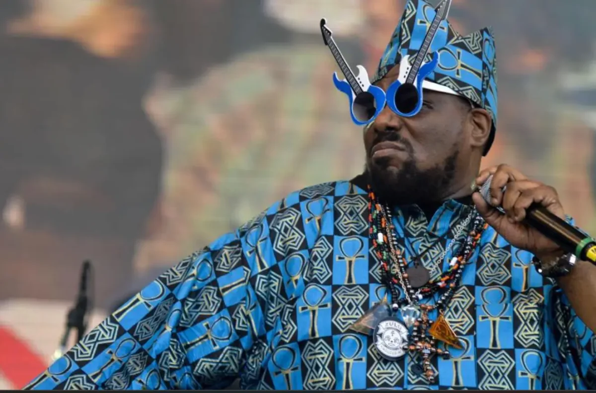 Afrika Bambaataa performing on stage wearing a blue patterned outfit and guitar-shaped sunglasses while holding a microphone